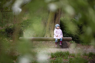 Little girl wearing blue hat and pink coat sitting on bench in a park - EYAF00189