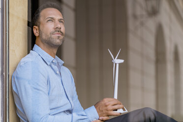 Businessman with wind turbine model behind windowpane thinking - DIGF06938