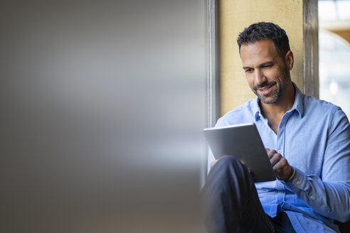 Lächelnder Geschäftsmann, der am Fenster sitzt und ein Tablet benutzt - DIGF06939