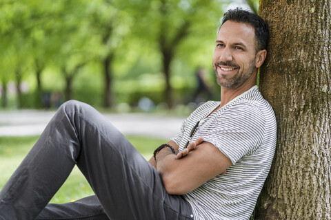 Lächelnder Mann lehnt an einem Baum im Park, lizenzfreies Stockfoto