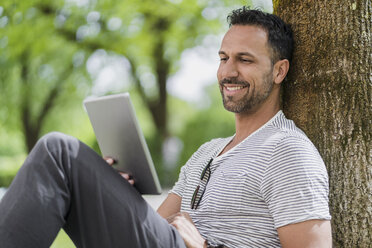 Lächelnder Mann lehnt an einem Baum im Park und benutzt ein Tablet - DIGF07005