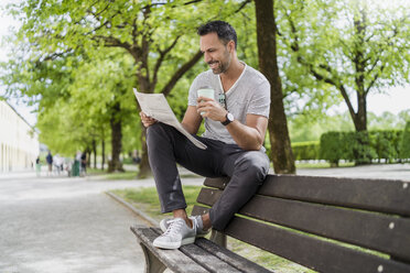 Smiling man with takeaway coffee sitting on park bench reading newspaper - DIGF07015