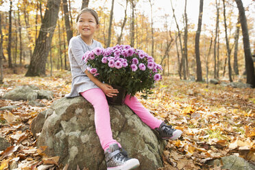 Lächelnde Mixed Race Mädchen sitzen auf einem Felsen im Herbst und halten Blumen - BLEF02720