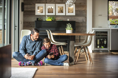 Lächelnder Vater und Sohn sitzen auf dem Boden und schreiben SMS auf ihren Handys - BLEF02741