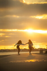 Mädchen hält sich an den Händen, während sie bei Sonnenuntergang mit dem Skateboard am Wasser fährt - BLEF02787