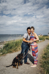 Porträt eines schwangeren lesbischen Paares mit Hund, das sich am Wasser umarmt - BLEF02842