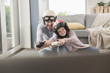 Young man and little girl wearing biker helmets, playing racing game with gaming consoles - UUF17342