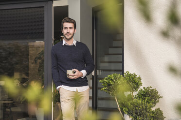 Young man leaning in door of his house, with a cup of coffee - UUF17373