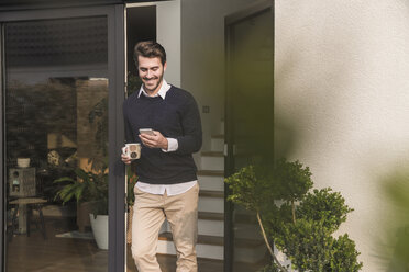 Young man leaning in door of his house, holding cup of coffee, using smartphone - UUF17374