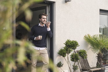 Young man leaning in door of his house, holding cup of coffee, using smartphone - UUF17376