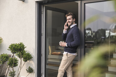 Young man leaning in door of his house, holding cup of coffee, using smartphone - UUF17377