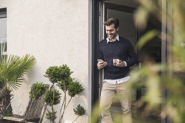 Young man leaning in door of his house, holding cup of coffee, using smartphone - UUF17379