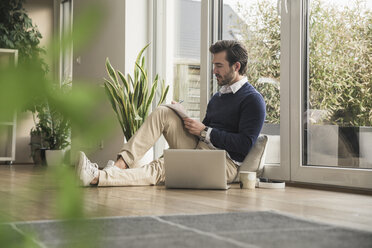 Junger Mann sitzt im Wohnzimmer, lehnt sich ans Fenster, arbeitet am Laptop und macht sich Notizen - UUF17380