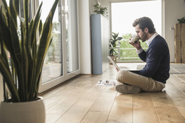 Young man sitting cross-legged in front of window, using laptop, drinking coffee - UUF17389