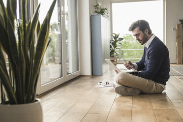 Young man sitting cross-legged in front of window, using laptop - UUF17391