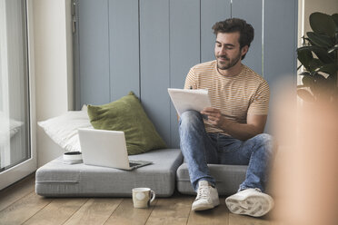 Young man sitting on mattress, using laptop, taking notes - UUF17392