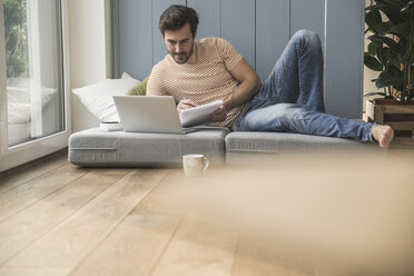 Young man sitting on mattress, using laptop, taking notes - UUF17394