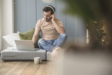 Young man sitting on mattress, using laptop with headphones - UUF17396