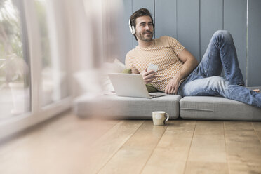 Young man sitting on mattress, using laptop with headphones - UUF17398