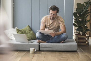 Young mamn sitting on mattress, taking notes, using headphones - UUF17399