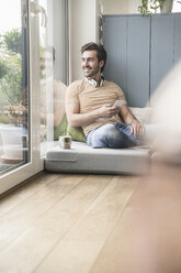 Young man sitting on mattress, using smartphone with headphones around neck - UUF17401