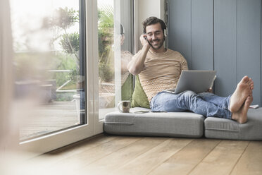 Young man sitting on mattress, using laptop with headphones - UUF17402