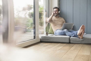 Young man sitting on mattress, using laptop with headphones - UUF17403