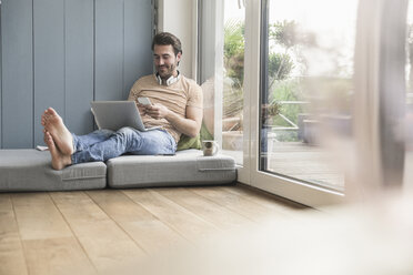 Young man sitting on mattress, using smartphone with headphones around neck - UUF17404