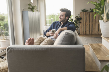 Young man sitting on couch at home, drinking coffee - UUF17406