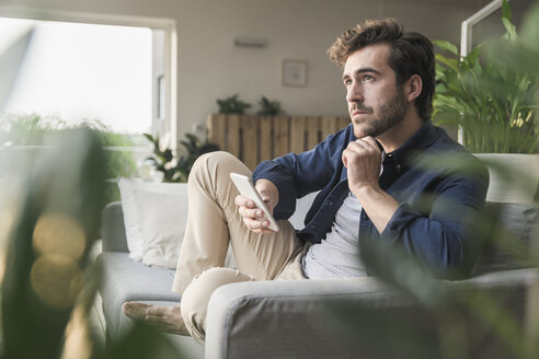 Junger Mann sitzt zu Hause auf der Couch und benutzt sein Smartphone - UUF17410