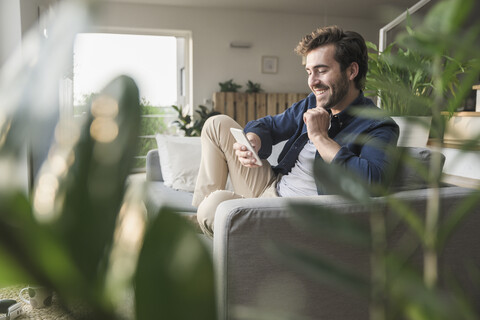 Junger Mann sitzt zu Hause auf der Couch und benutzt sein Smartphone, lizenzfreies Stockfoto