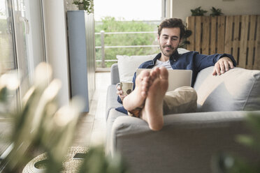 Young an sitting on couch, using laptop, drinking coffee - UUF17414