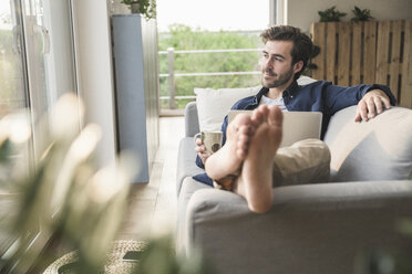 Junger Mann sitzt auf der Couch, benutzt Laptop, trinkt Kaffee - UUF17415