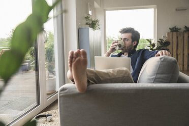 Young an sitting on couch, using laptop, drinking coffee - UUF17416