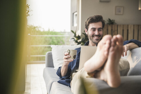 Young an sitting on couch, using laptop, drinking coffee - UUF17417