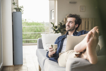 Young an sitting on couch, using laptop, drinking coffee - UUF17418