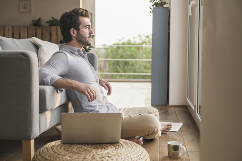 Junger Mann sitzt zu Hause auf dem Boden, benutzt einen Laptop und schaut aus dem Fenster, lizenzfreies Stockfoto