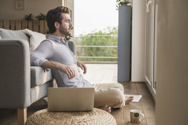 Young man sitting at home on floor, using laptop, looking out of window - UUF17420