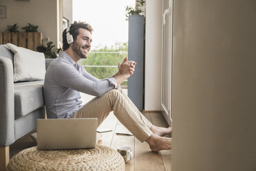 Young man sitting at home on floor, using laptop, listening music - UUF17422