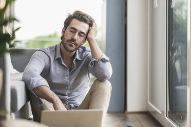 Young man sitting at home on floor, using laptop, taking a break - UUF17429