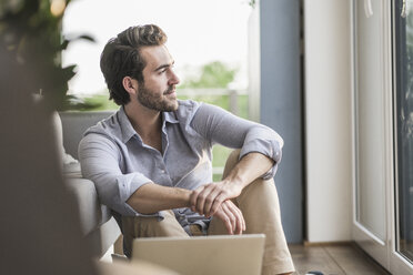 Young man sitting at home on floor, using laptop, looking out of window - UUF17430