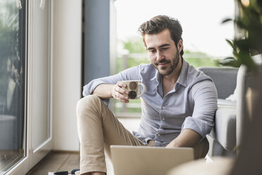 Young man sitting at home on floor, using laptop, drinking coffee - UUF17431