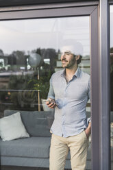 Young man looking out of window, using smartphone - UUF17438