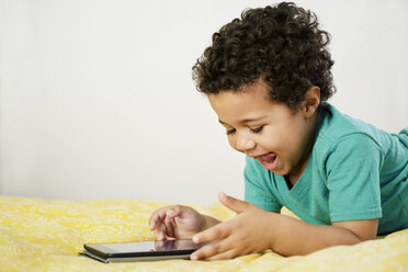 Aufgeregter Mixed-Race-Junge, der auf dem Bett liegt und ein digitales Tablet benutzt - BLEF02940