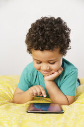 Lächelnder Mixed-Race-Junge, der auf dem Bett liegt und ein digitales Tablet benutzt - BLEF02941