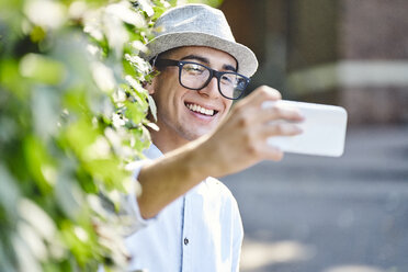 Porträt eines jungen glücklichen Mannes, der ein Selfie mit einer Hecke im Hintergrund macht - BSZF01063