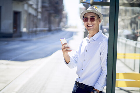 Glücklicher junger Mann mit Mobiltelefon an einer Straßenbahnhaltestelle in der Stadt, lizenzfreies Stockfoto