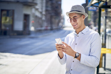 Glücklicher junger Mann, der an einer Straßenbahnhaltestelle in der Stadt sein Telefon überprüft - BSZF01069