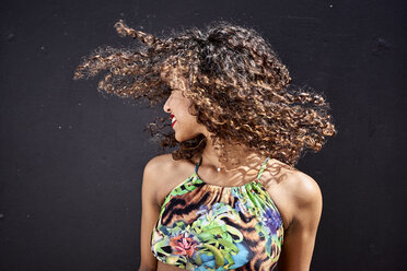 Smiling young woman tossing her curly hair in front of dark background - VEGF00141