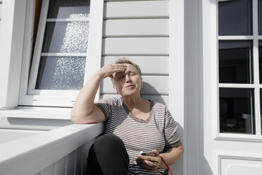 Relaxed senior woman sitting on porch in sunshine - KMKF00915